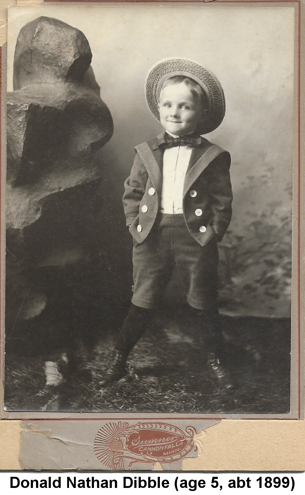IMAGE/PHOTO: Donald Nathan Dibble (age 5, abt 1899): Black and white studio photo of a small boy wearing Little Lord Fauntleroy suit with straw hat cocked back on his head, bow tie, knickers and knee sox, standing next to a large rock in front of a cloth backdrop with his feet wide apart, knees turned in, hands in pockets, and his eyes aimed to the right.
